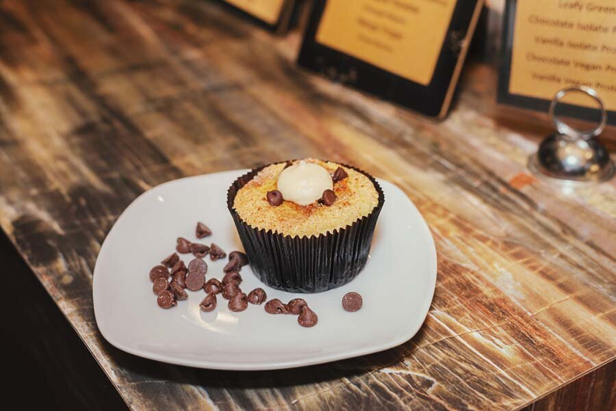 A chocolate muffin topped with sliced almonds on a white plate, surrounded by scattered chocolate chips, on a wooden table with menu stands in the background.
