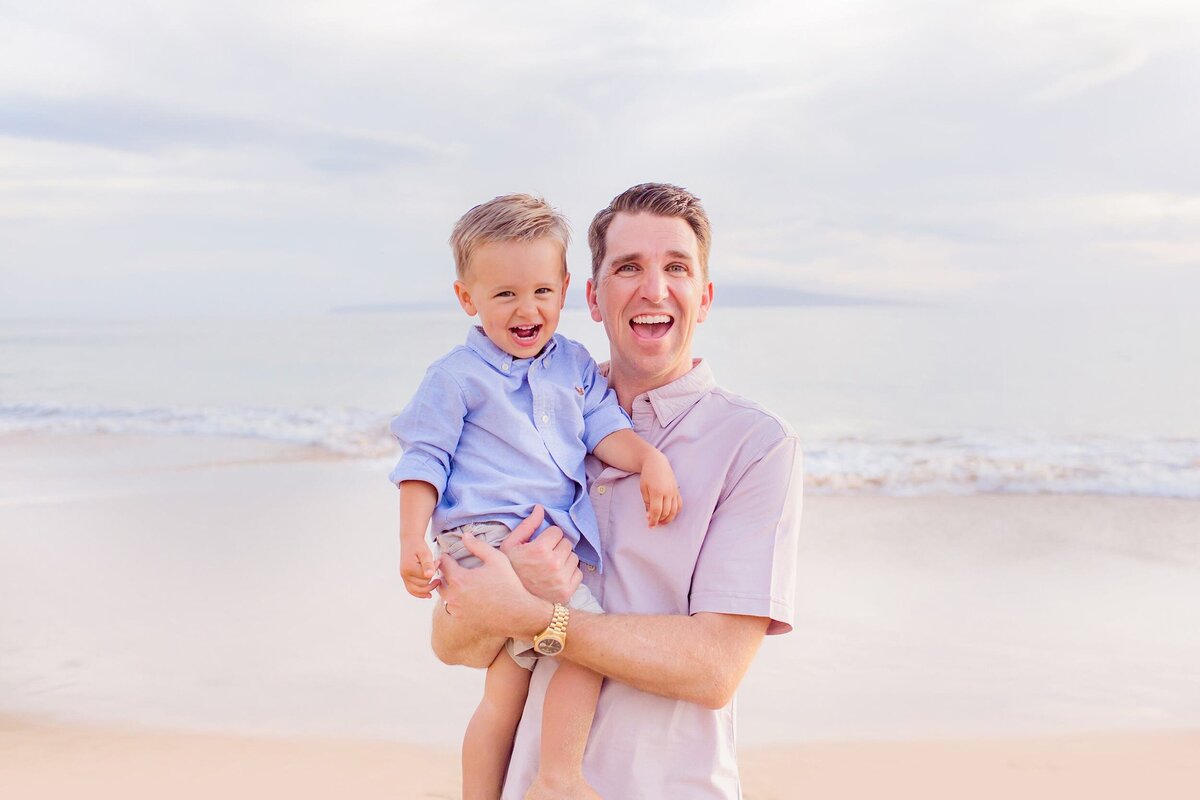 Dad and young son wear button up shirts and smile at the camera on the beach in Wailea