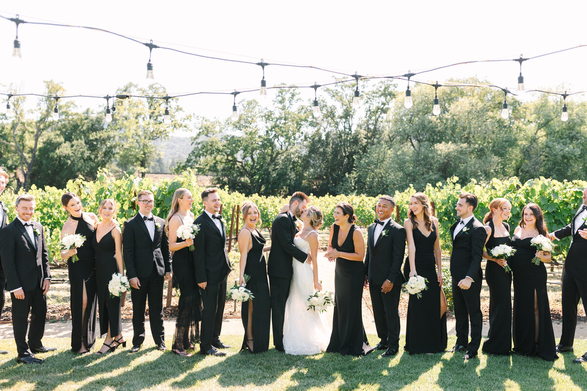 bride and groom with bridal party dressed in black standing in napa vineyard underneath twinkly lights.