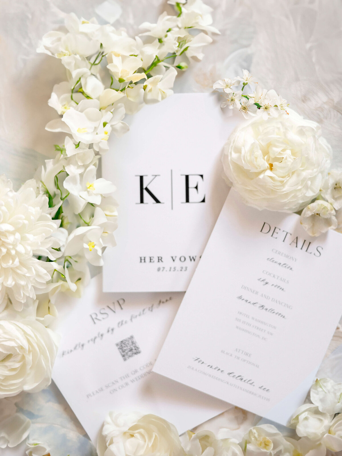 A flat lay of elegant white wedding stationery surrounded by white flowers. The cards include initials K | E, details, and an RSVP with a QR code, set on a soft, light fabric background.