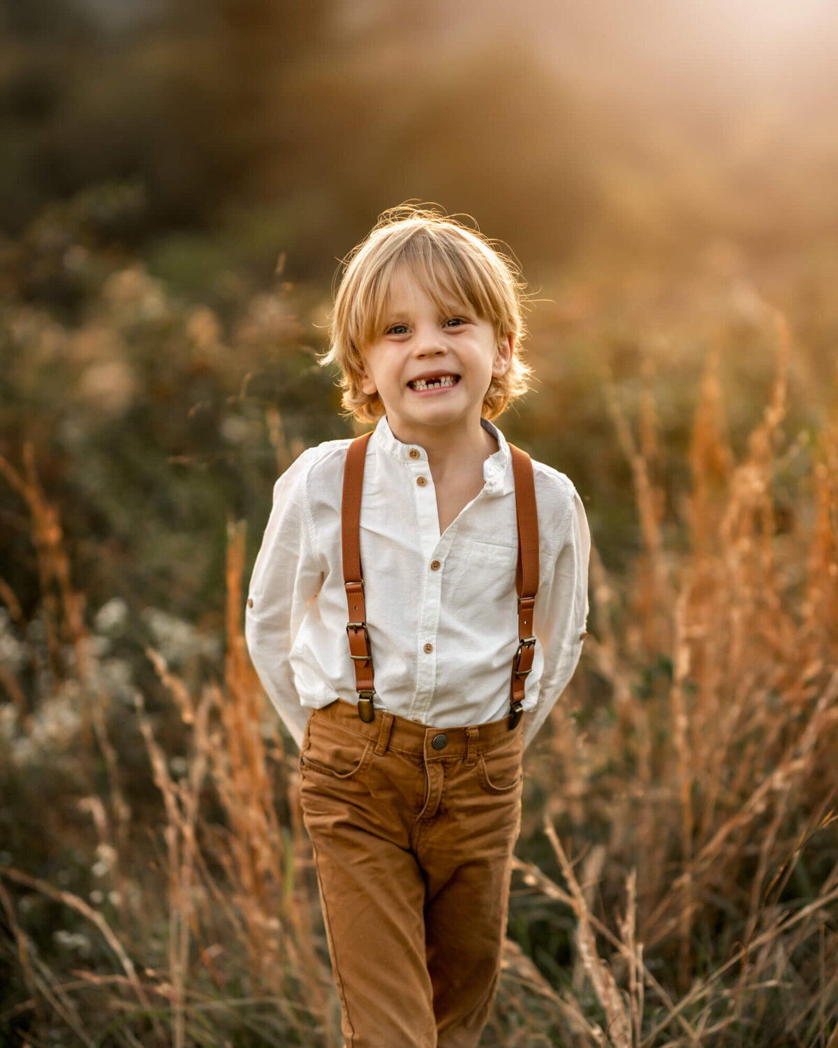 A adorable boy with missing front teeth and suspenders standing in the tall grass at a park in Asheville