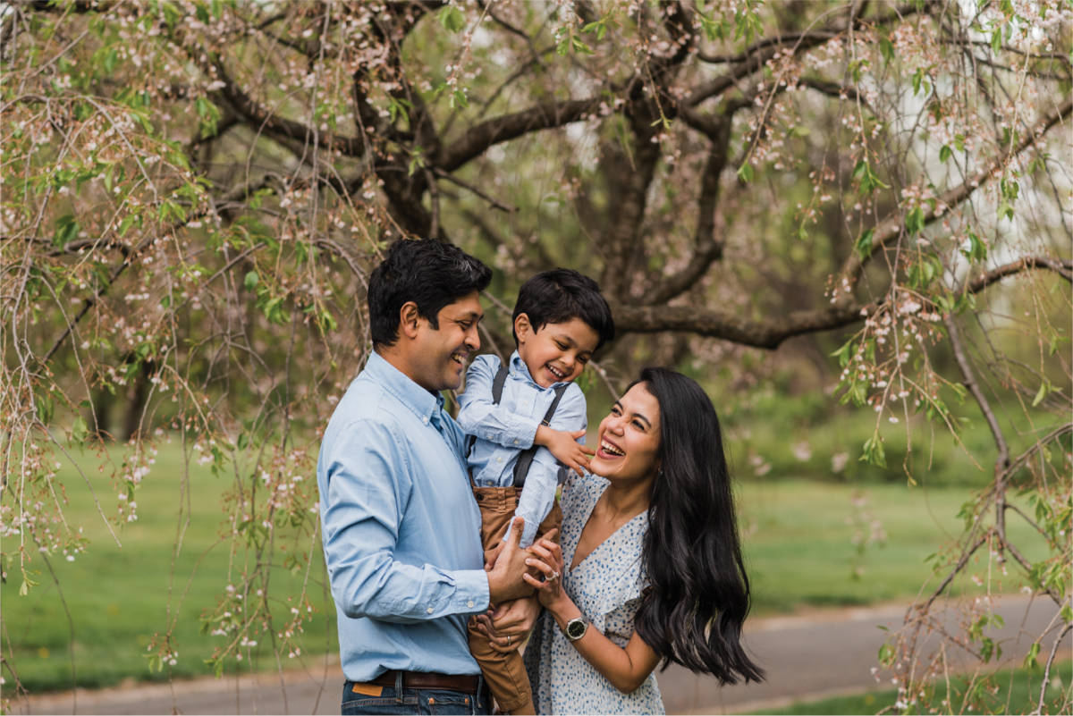 A couple smiling and carrying a young boy under a large tree with flowers.