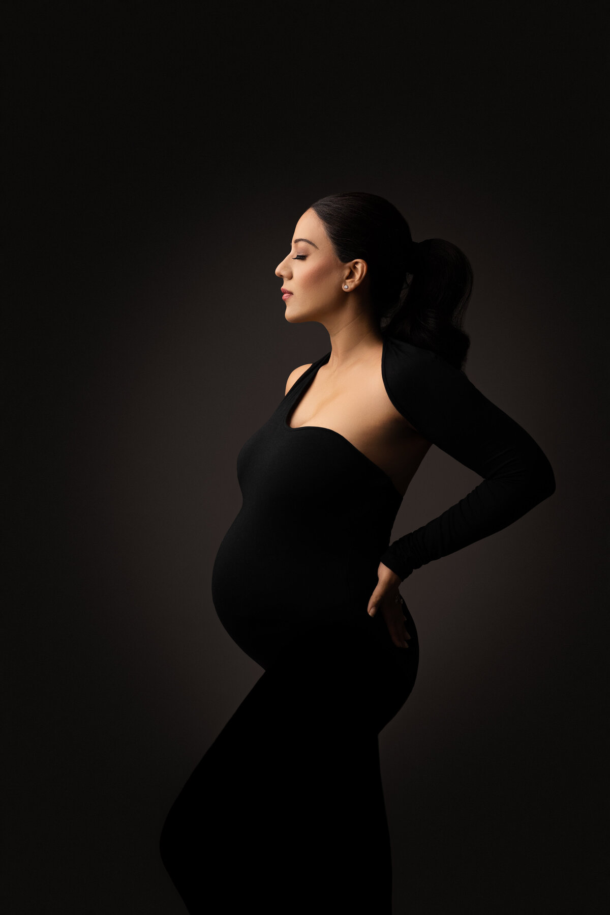 In this artistic, fine art photo captured by New Jersey's best maternity photographer Katie Marshall, an expectant mom is standing side-profile to the camera. The woman is in a luxurious maternity one-sleeve body-con dress is standing side profile to the camera. Her hands are resting on the small of her back.  The woman's eyes are closed and  featuring the side profile of her face.