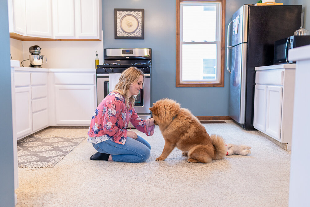 A woman in a floral top kneels on a kitchen floor, interacting with a fluffy, medium-sized dog. the room has white cabinets and a black refrigerator.