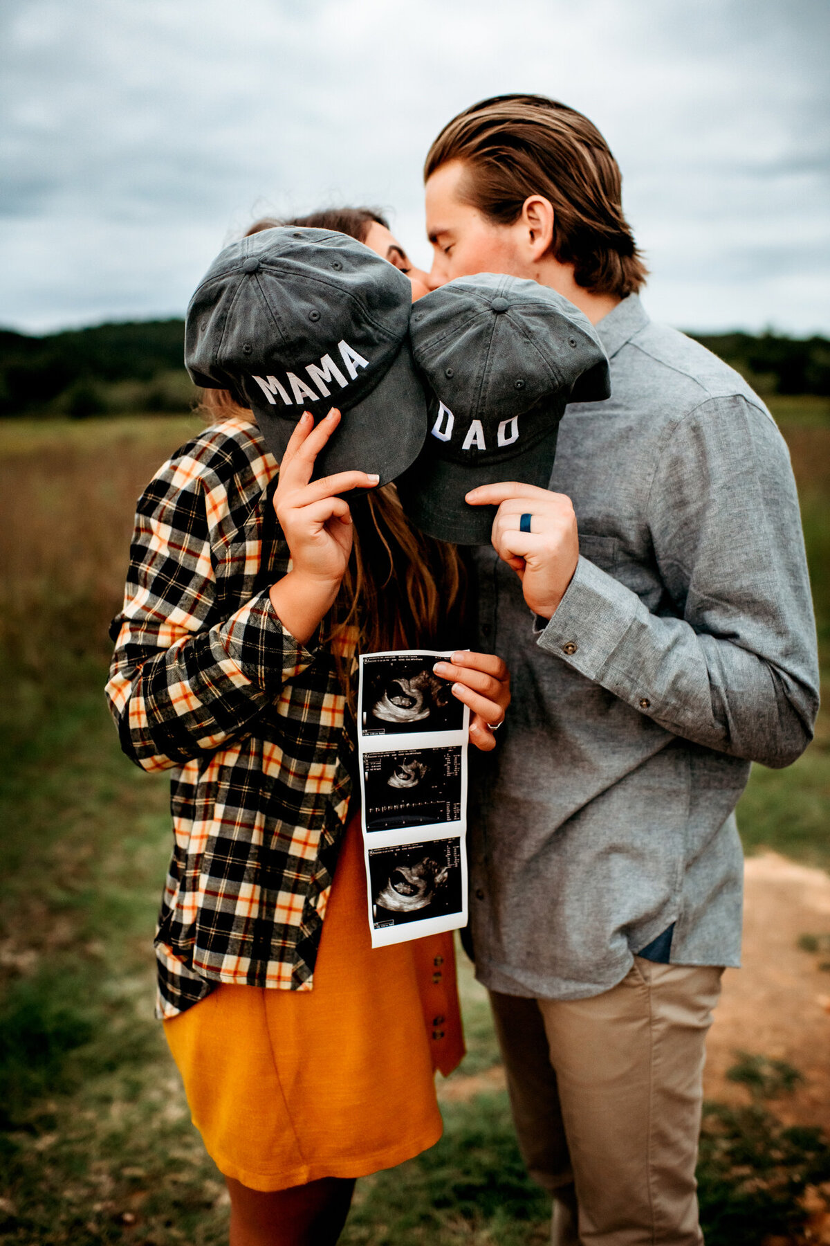 Couples Photography, Woman holding hat that says "Mama" and also holding an ultrasound, hides her face behind the hat as she kisses a man that holds a hat that says "Dad" and kisses the hat.