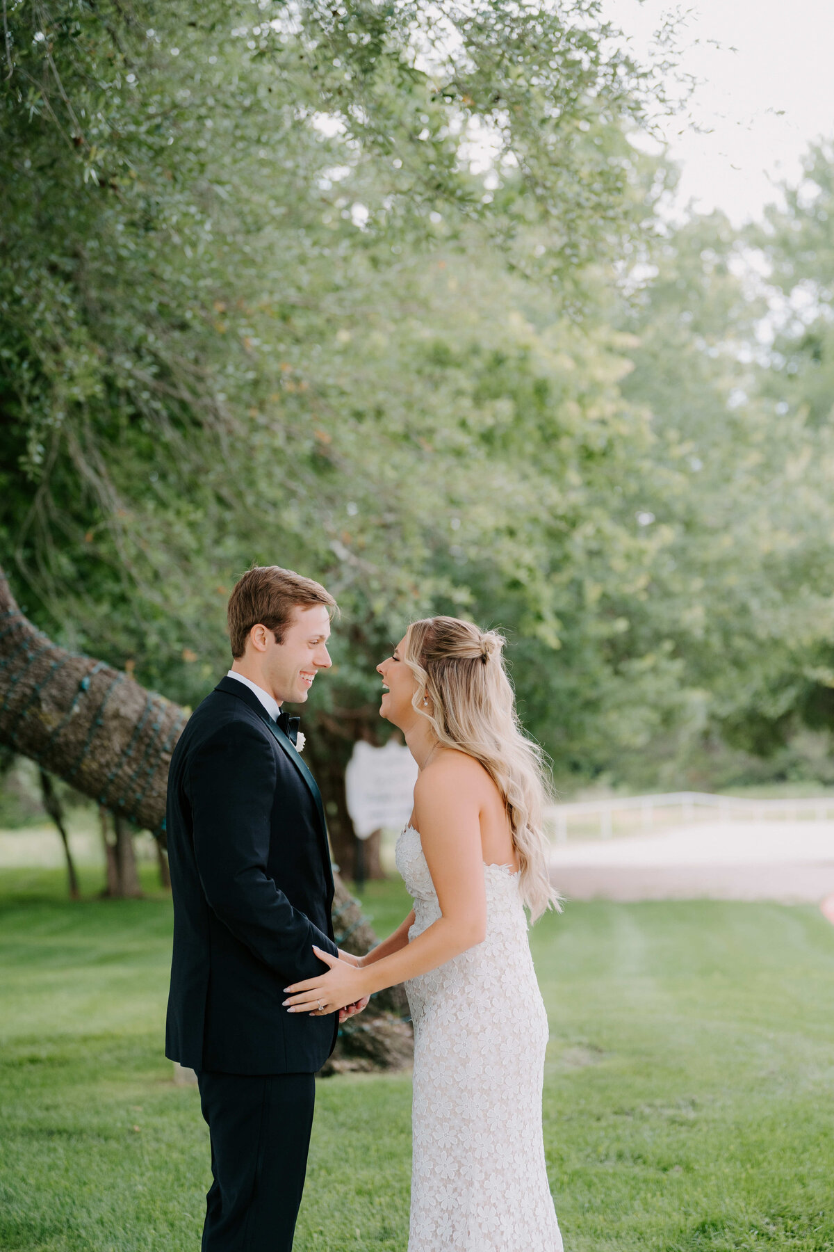 Bride and groom share a joyful moment outdoors under a large tree, surrounded by lush greenery. The scene conveys happiness and love.