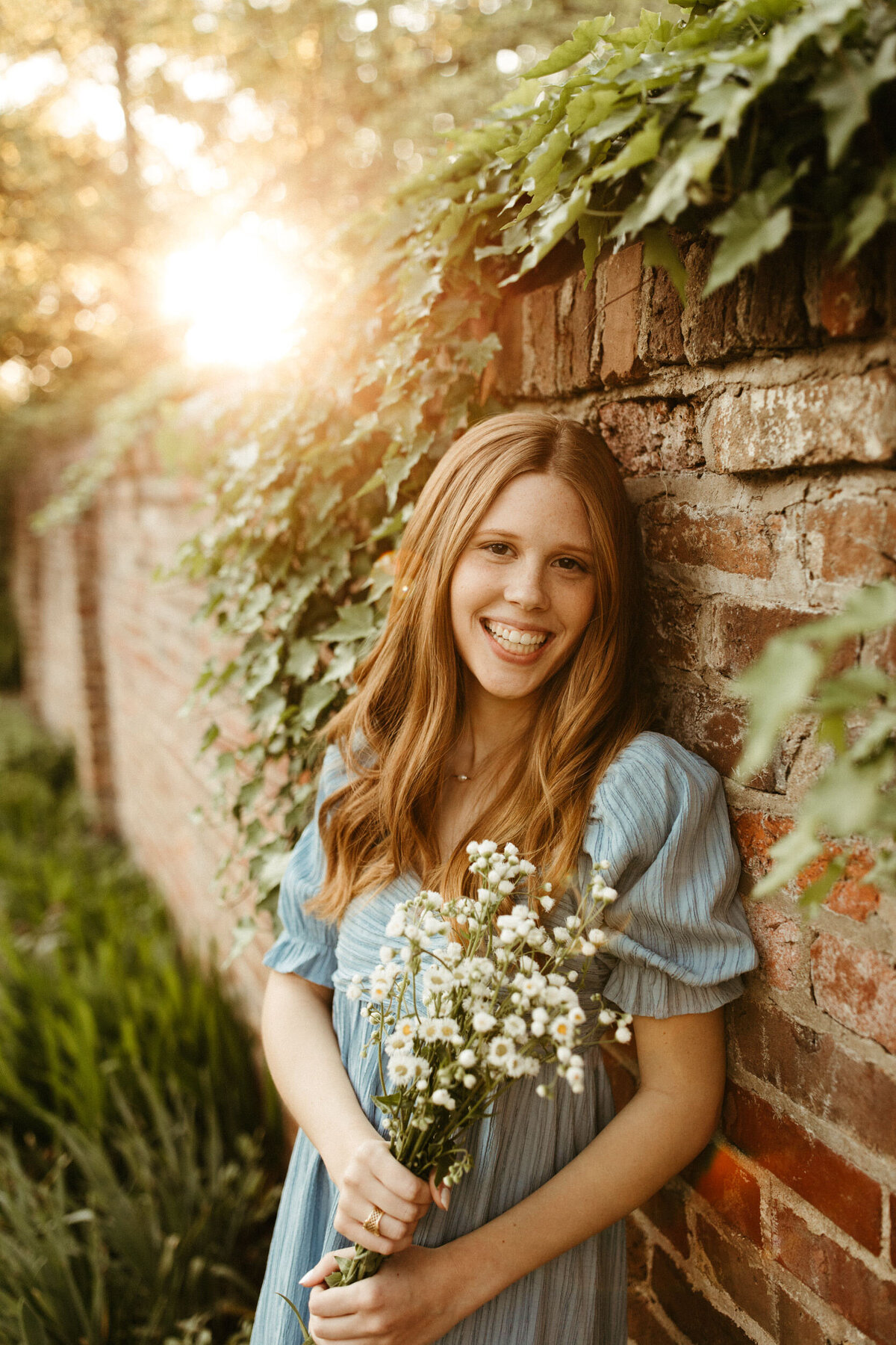 Girl senior holding a bundle of wildflowers and leaning against a brick wall with ivy growing along it