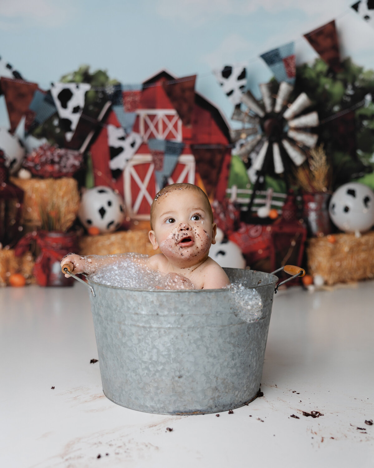 Little boy sitting in a bubbly metal tub with cake still on his face, in front of a rustic barn-themed backdrop.
Photograph by Yucaipa cake smash photographer Kaitlyn Dawn Photography.