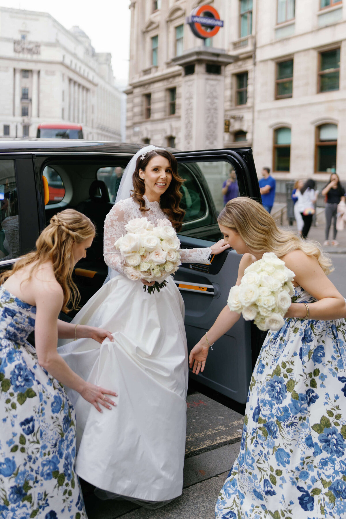bridesmaids in blue patterned dresses help the bride exit her cab as she arrives at her chic city wedding ceremony in a traditional london black cab