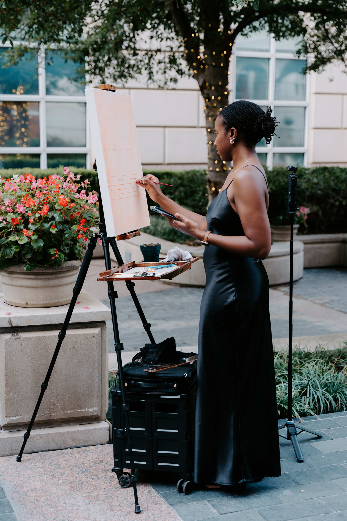Woman in a black dress painting on an easel outdoors, surrounded by flowers and fairy-lit trees. The atmosphere is serene and focused.