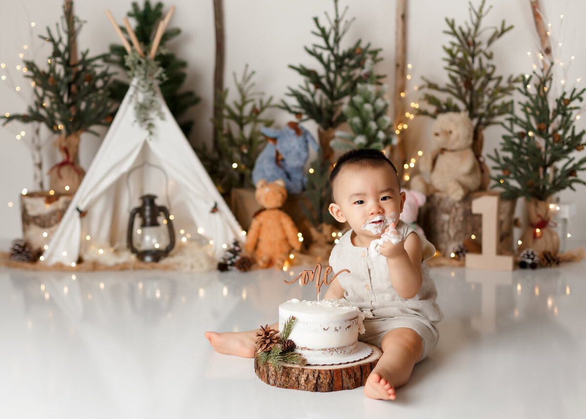 Rustic Winnie the Pooh themed cake smash in West Palm Beach, FL newborn and cake smash photography studio.  Baby boy wearing white is sitting with white cake between his legs with a fist full of cake looking at the camera. In the background, there are green evergreen trees, a canvas teepee tent, and vintage Winnie the Pooh stuffed characters.