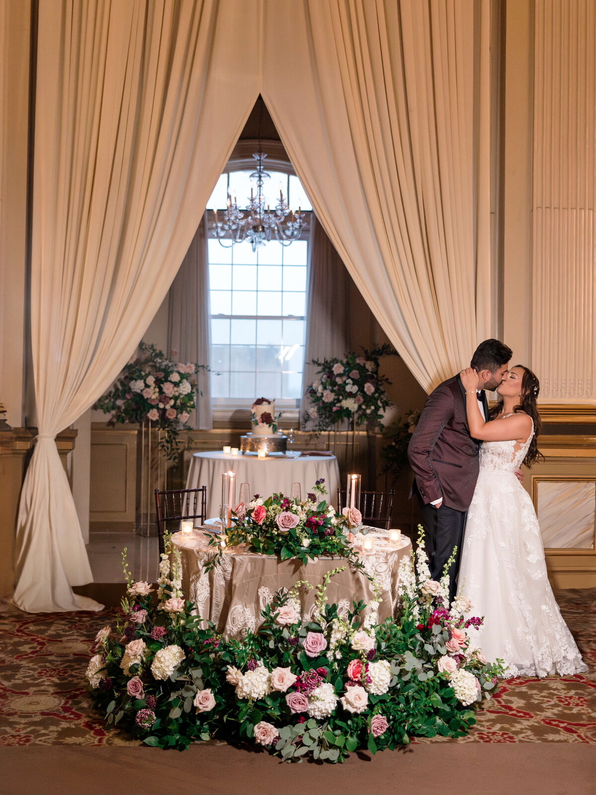 A couple shares a kiss in an elegant room adorned with floral arrangements. They stand beside a table with candles and flowers, framed by large curtains. A chandelier hangs overhead, adding a touch of opulence to the setting.