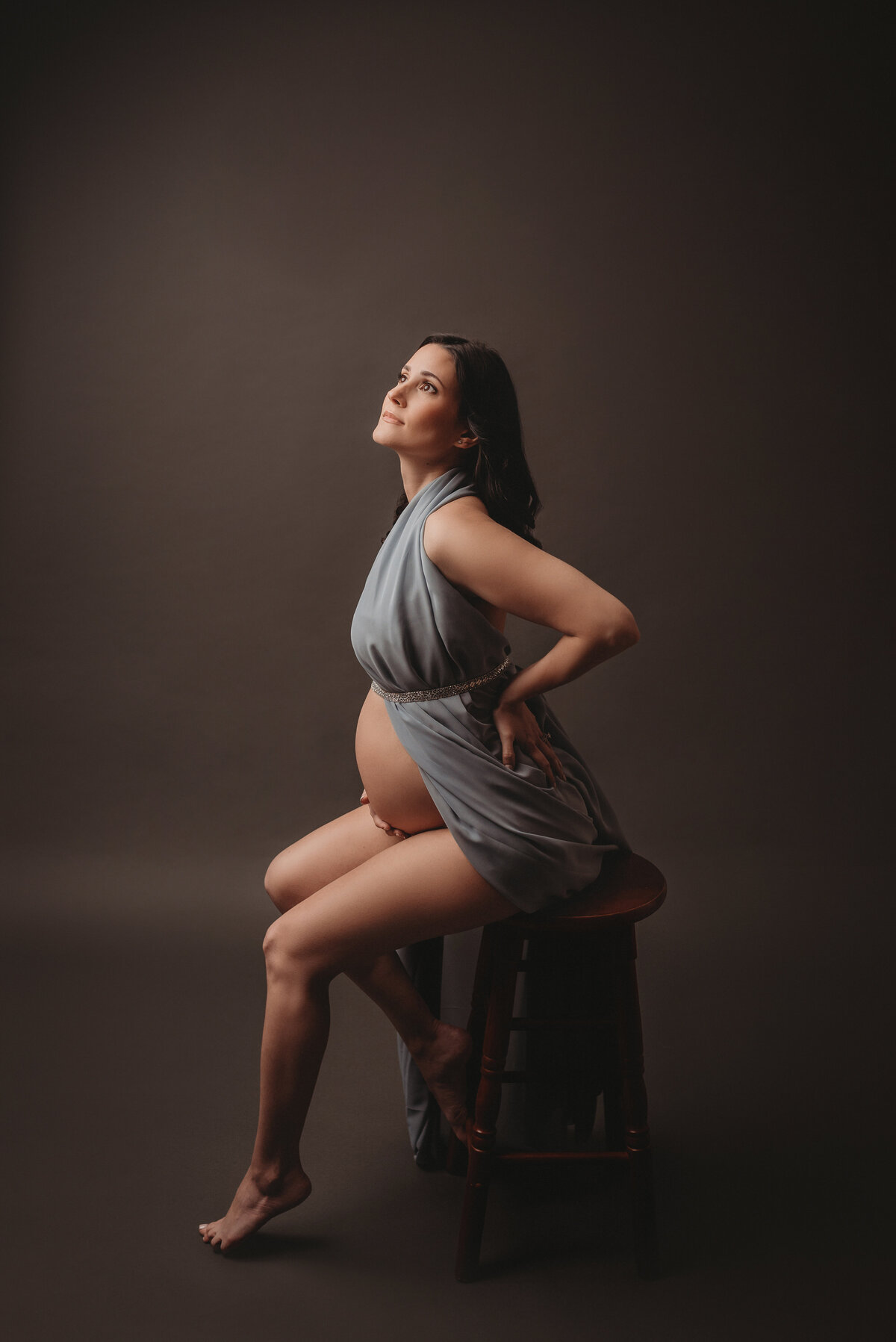 Pregnant mom to be sitting on stool with one leg up on rung of stool, hand on back and leaning forward looking up towards the light
