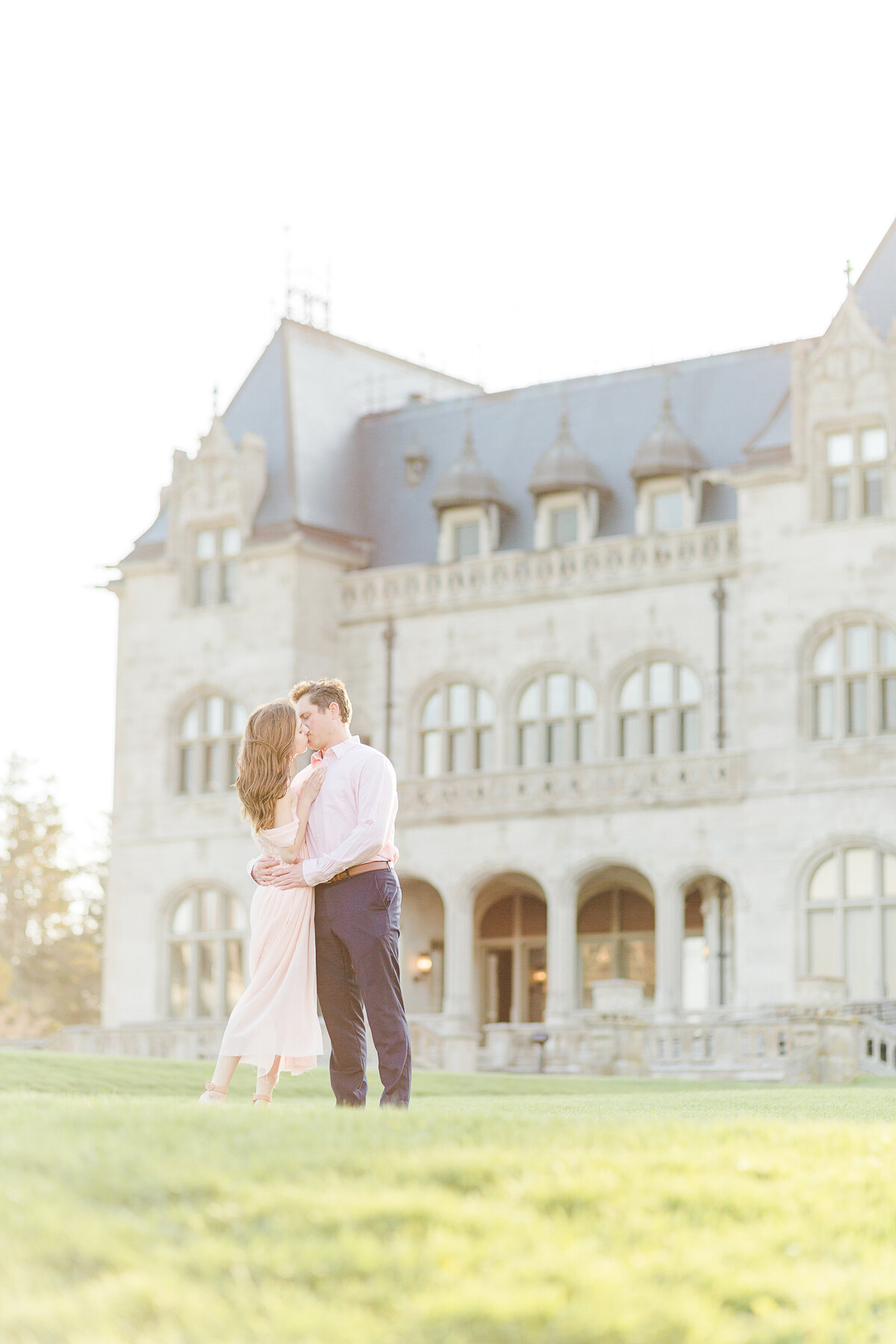 Couple stands in front of Salve Regina stone buildings in a warm embrace for their Newport, RI engagement session. They share a gentle kiss. The buildings are featured prominently in the background. Captured by best RI wedding photographer Lia Rose Weddings