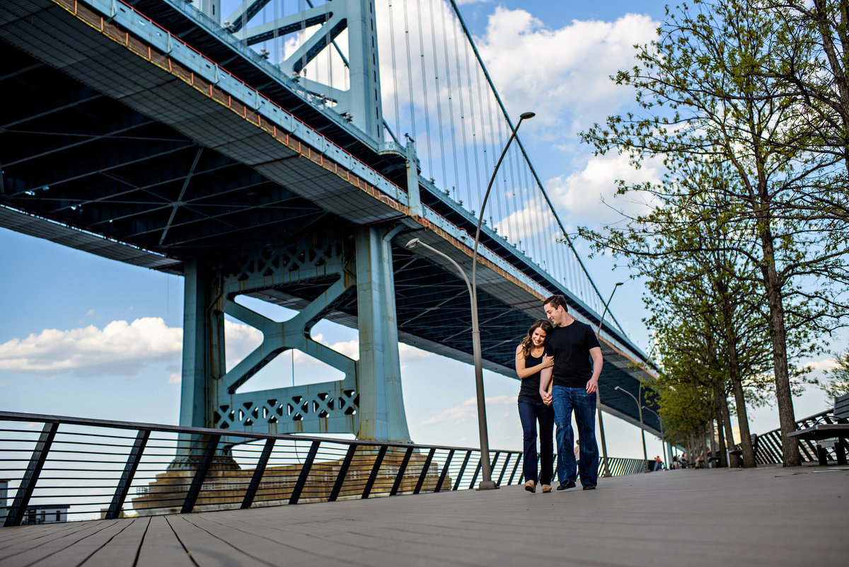 A happy couple walk along race street pier with ben franklin bridge behind them.