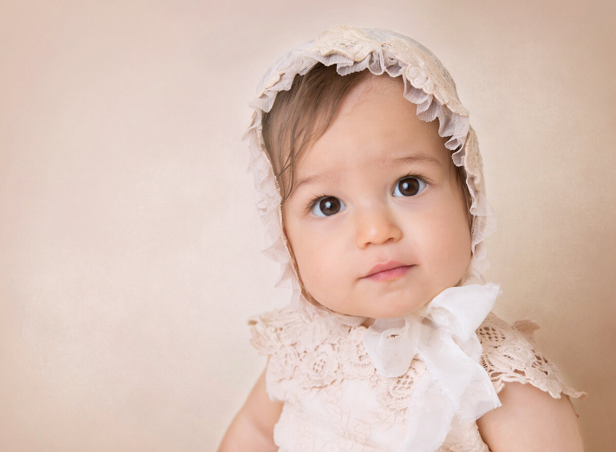 Baby girl looking at the camera intently during a 6-month baby milestone photoshoot. She is waring a lace outfit and matching lace bonnet.