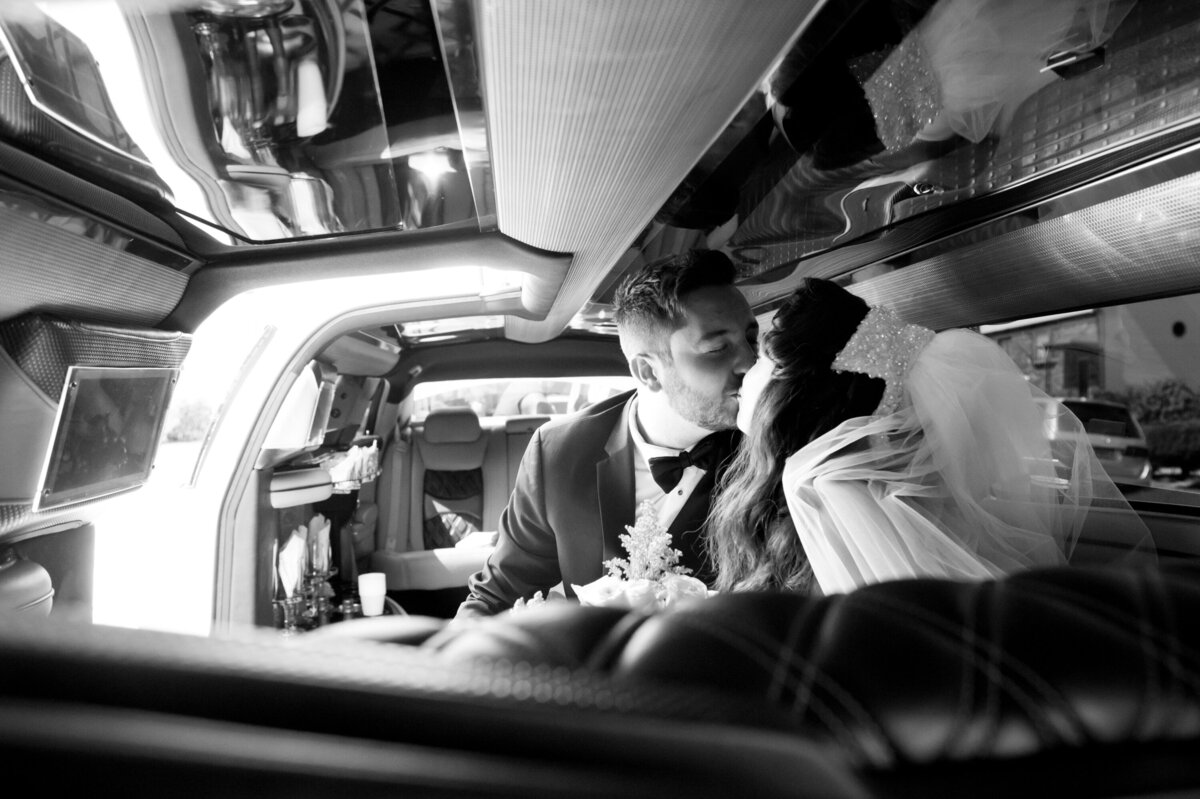 Black and white photo of a man in a suit with black bow kisses his bride in a limo Venetian New York wedding photographer Milestone Images