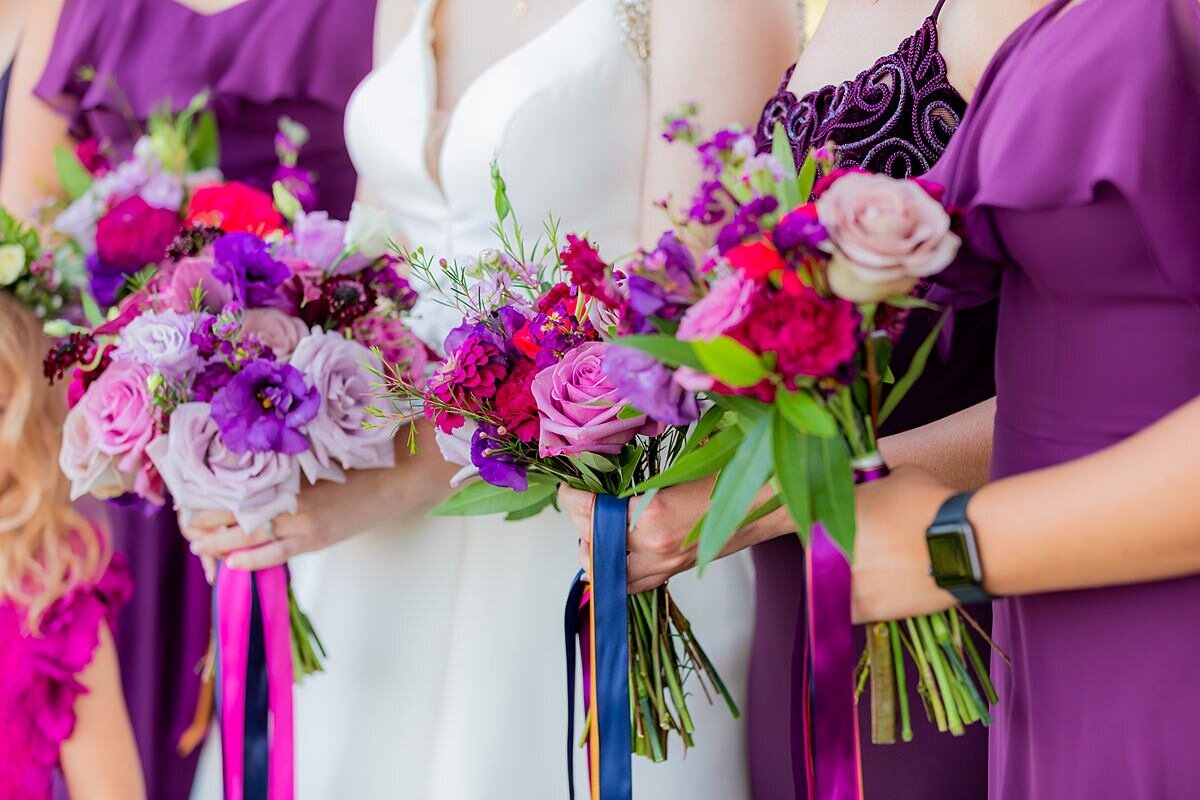 Bride and bridesmaids in purple dresses hold bouquets of pink roses, purple roses, red roses, greenery, pink and purple flowers with long streaming ribbons in royal blue and hot pink.