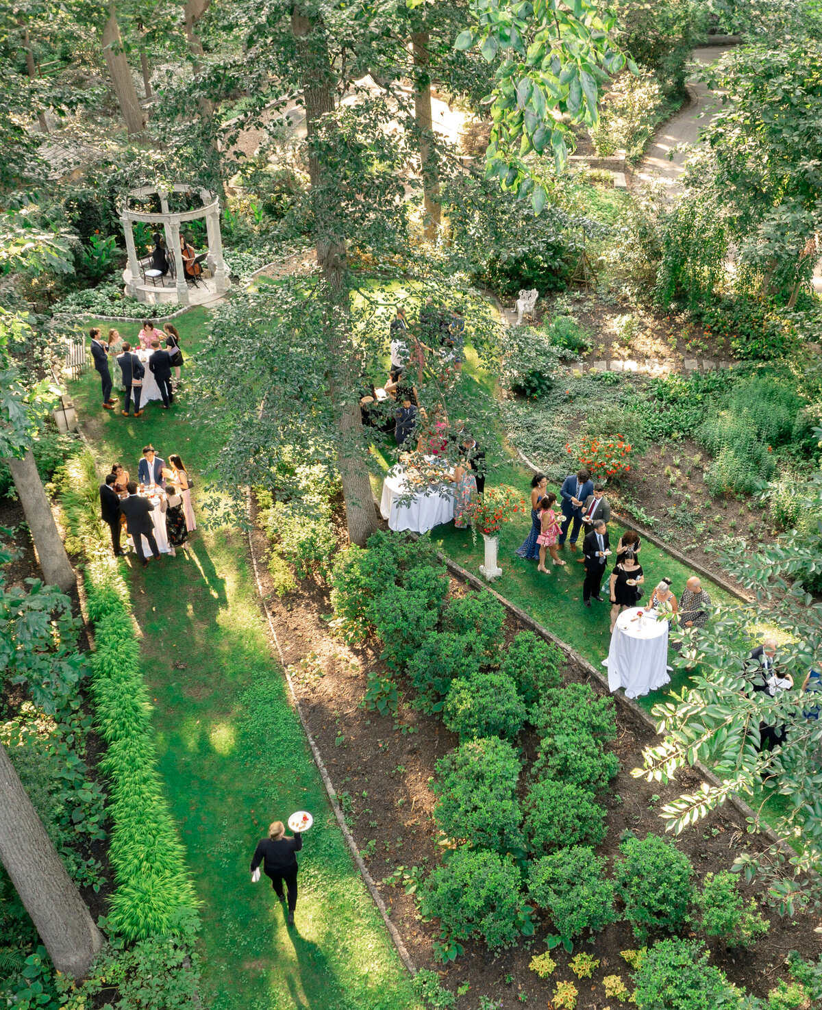 Aerial view of a lush garden with guests mingling at a formal outdoor event. People are gathered near tables set amidst vibrant greenery and trees. A white gazebo is visible in the background, enhancing the elegant atmosphere.
