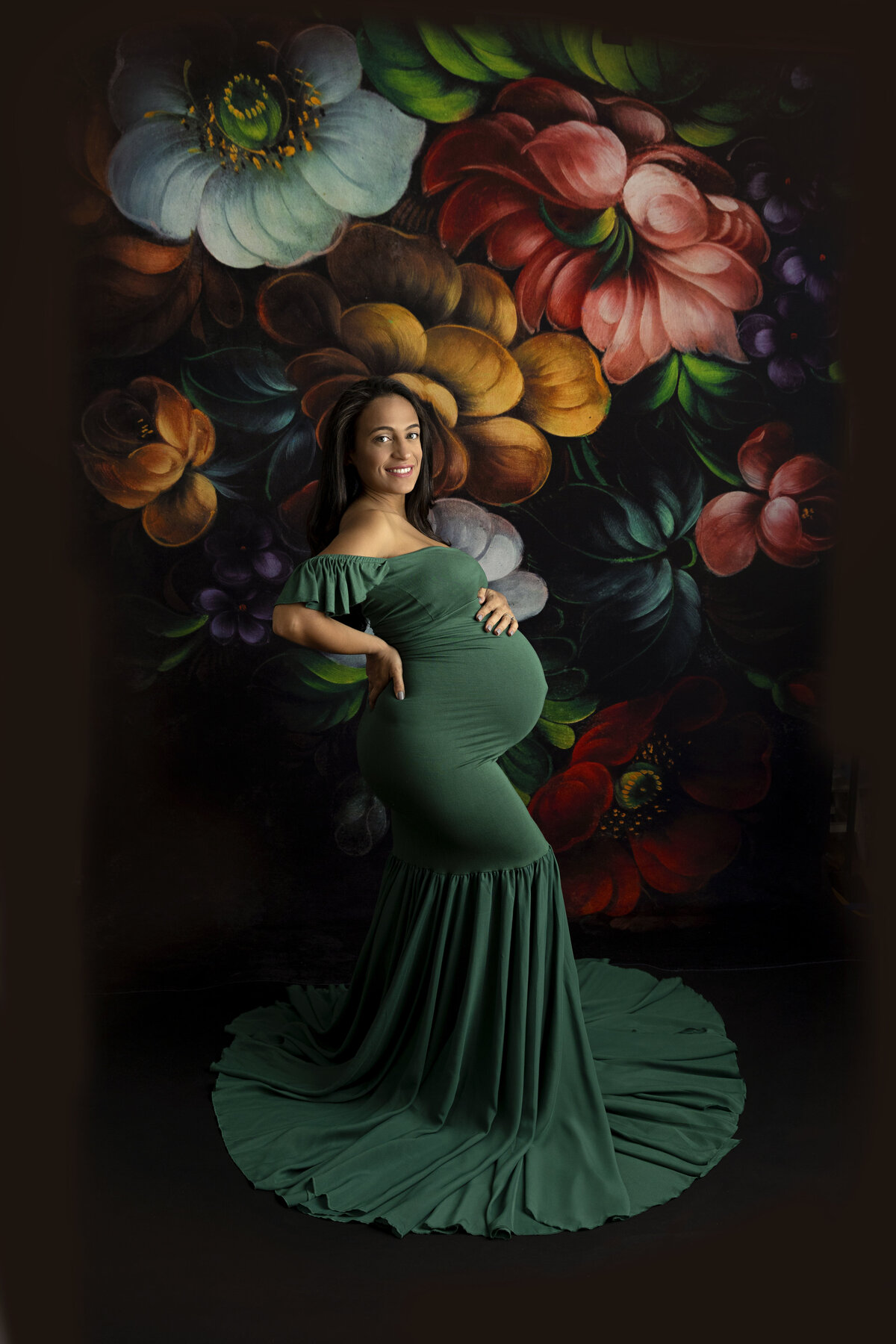 A mother to be in a green maternity gown smiles while standing in a studio with a floral print backdrop