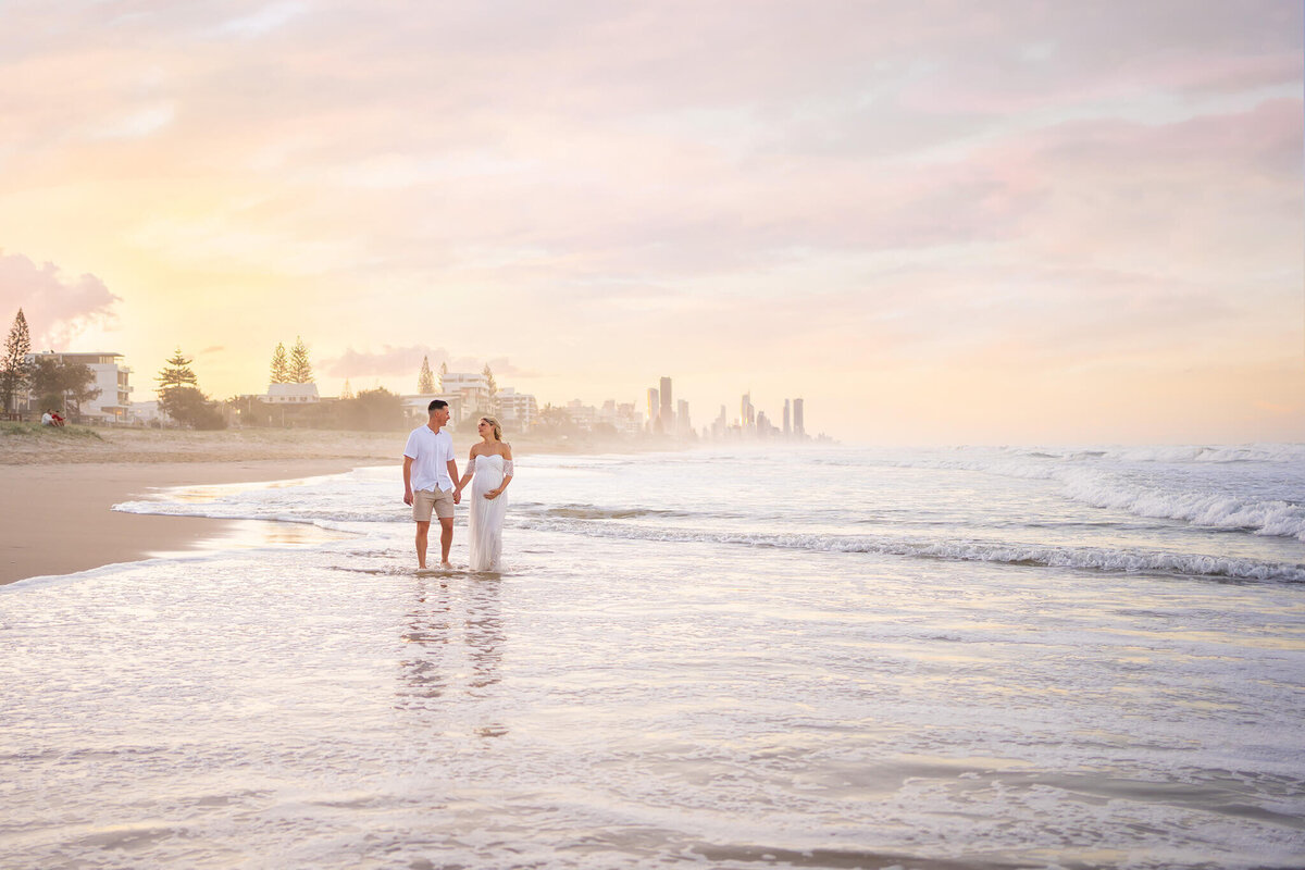 couple walking in seawater in gold coast with pastel sunset for maternity photos