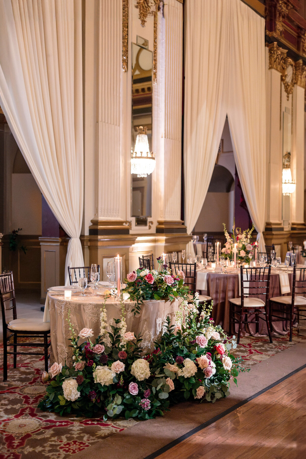 Elegant wedding reception with a round table draped in white cloth adorned with candles and glassware. A lavish floral arrangement with pink and white flowers is in front. Tall white curtains and ornate chandeliers decorate the opulent hall.