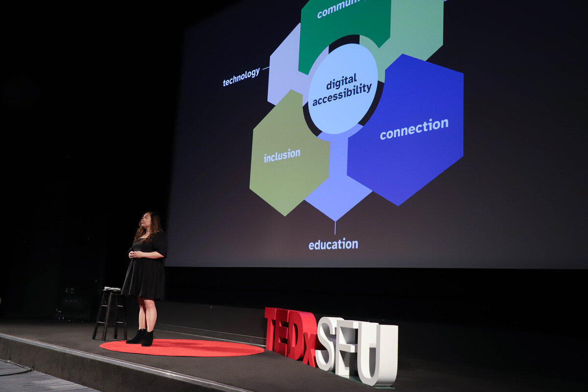 Anne stands on the TEDxSFU stage with the full graphic  behind her relating digital accessibility to community, connection, and inclusion