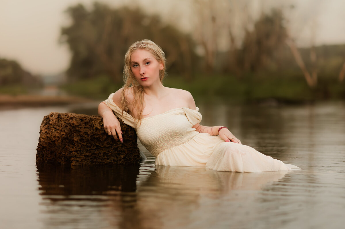 Female Roseville high school senior laying down on her side in the river leaning against a rock