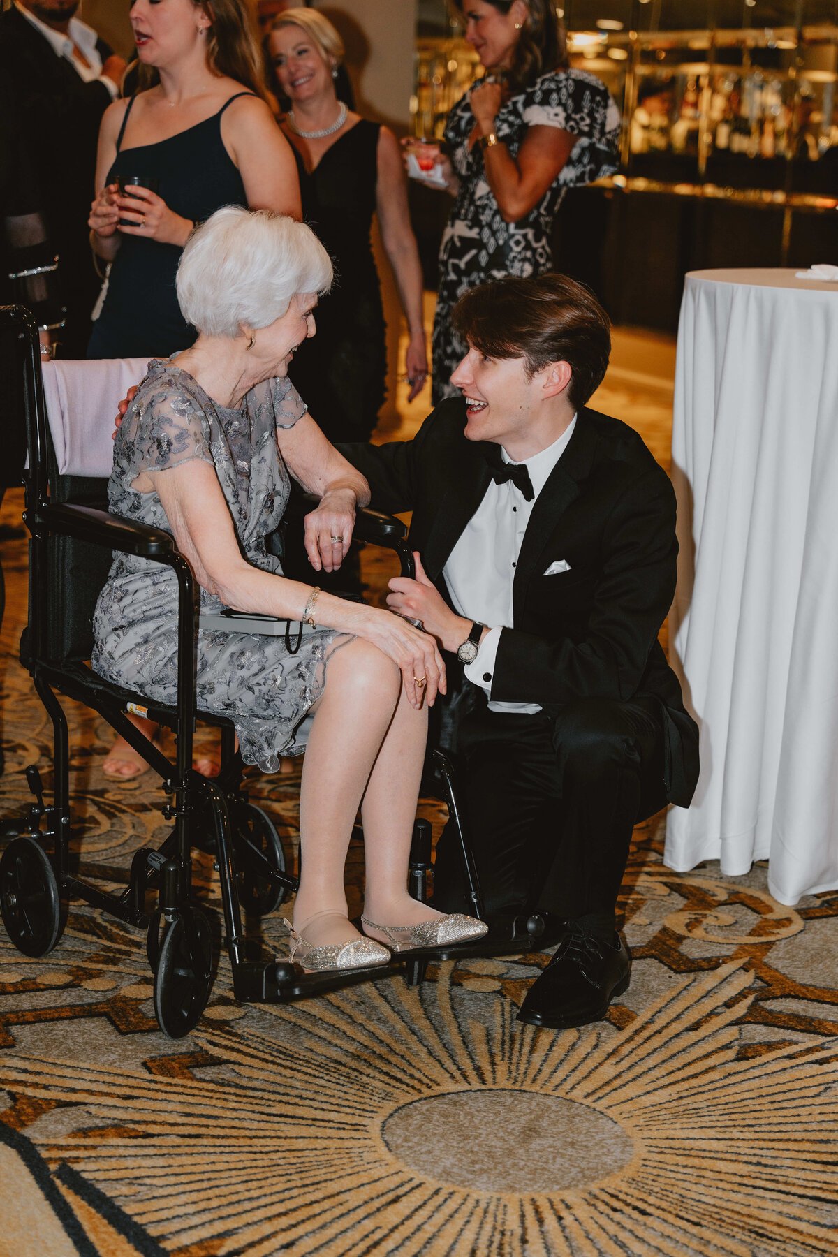 A young man in a tuxedo kneels beside an elderly woman in a wheelchair, both smiling warmly at each other. Elegant event setting with guests in the background.