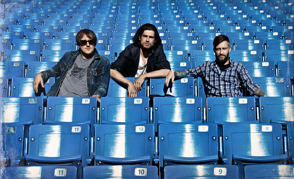 Musical trio portrait The Manvils sitting together amidst empty blue stadium seating