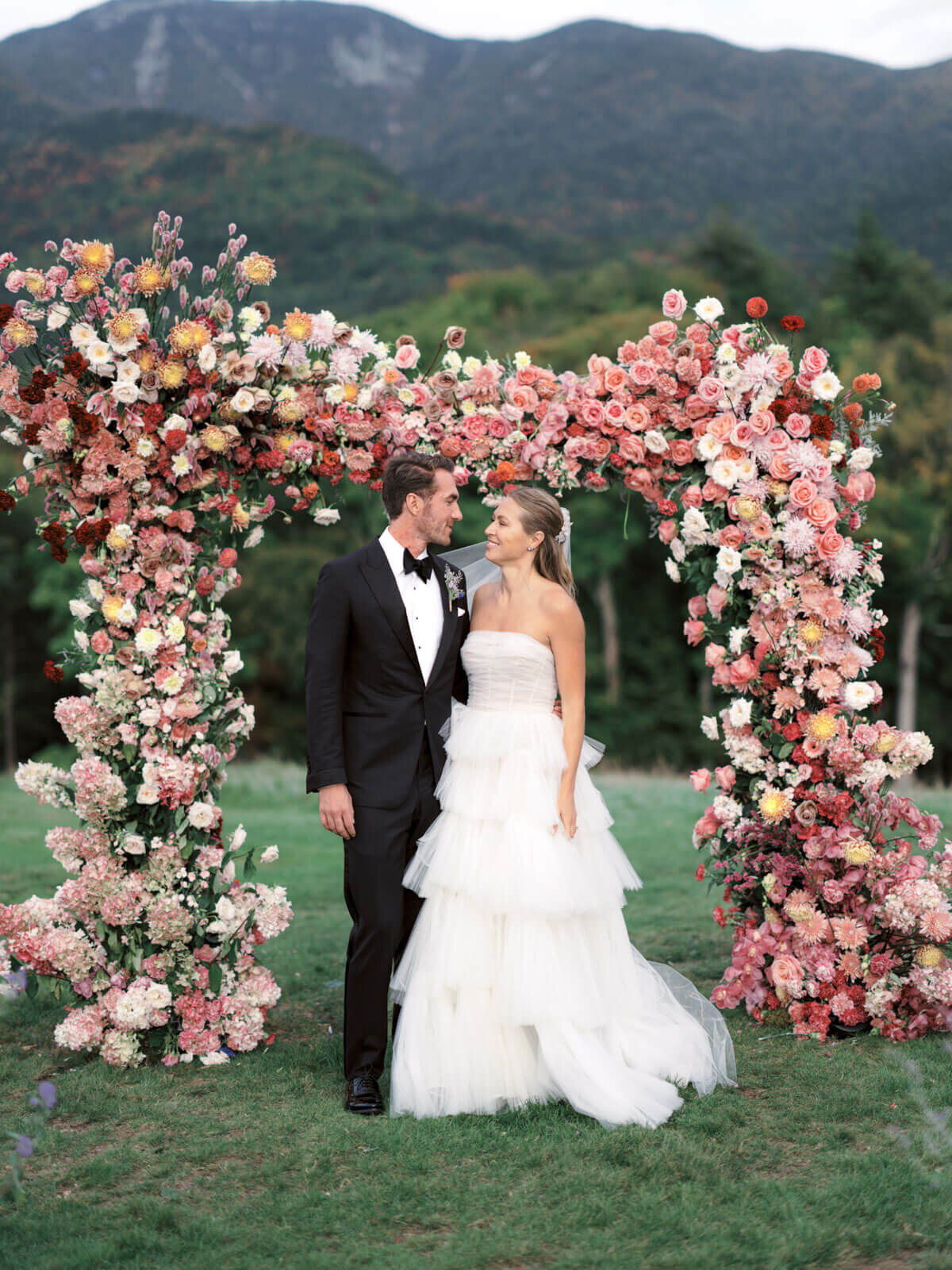 The bride and the groom are smiling at each other, with an arch full of flowers and mountains in the background at The Ausable Club, NY.