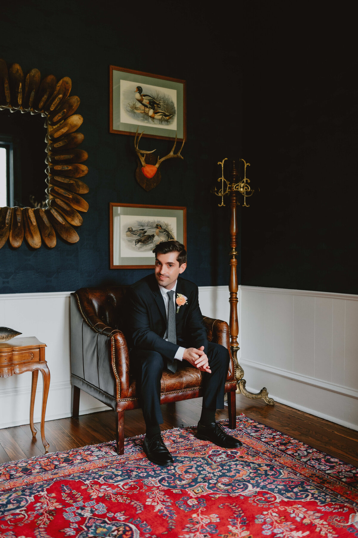 Groom in a black suit sitting on a leather chair in a stylish room with dark walls, vintage decor, and a patterned rug