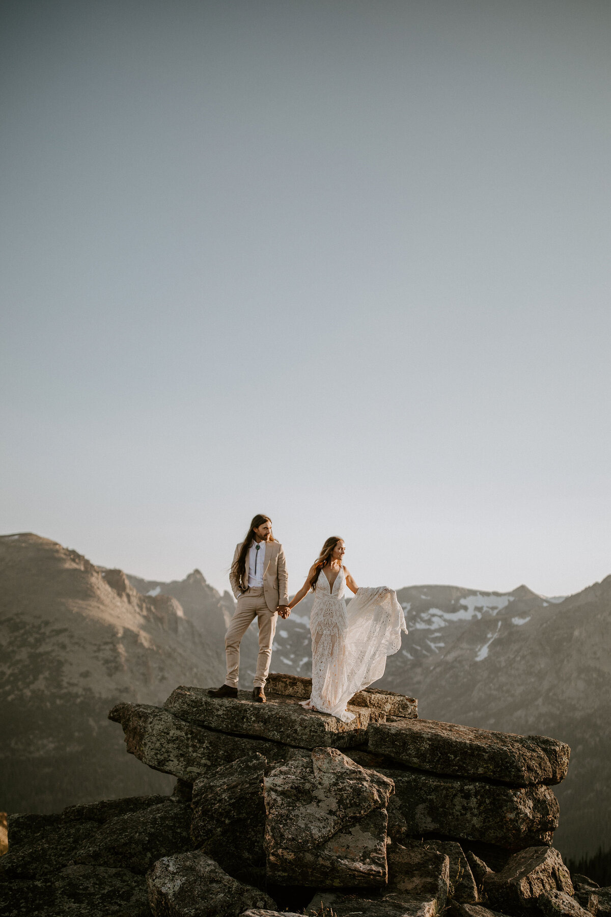 Bride and groom wearing an ivory suit and white wedding gown standing atop a rocky mountain landscape.