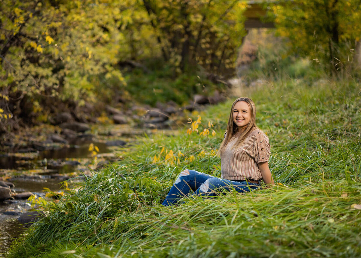 high school senior girl sitting in a grassy field near a shallow creek