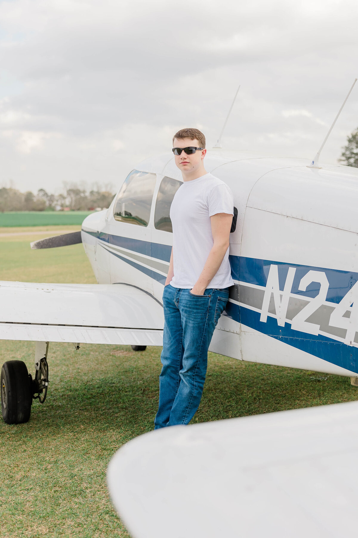 A person wearing sunglasses and a white t-shirt stands with hands in pockets, leaning against a small airplane with a blue stripe and tail number N24. The aircraft is parked on grass under a cloudy sky.