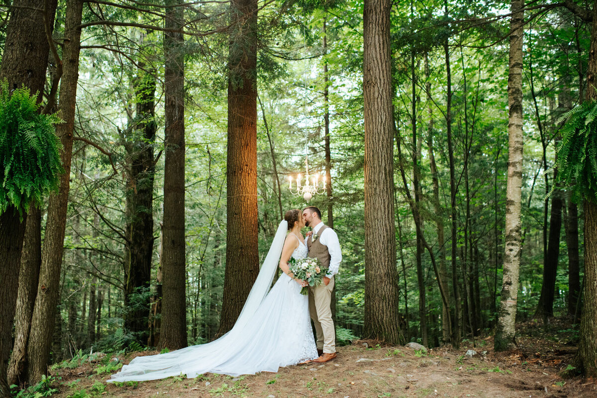 bride and groom at outdoorsy wedding kissing in forest under chandelier vermont adventure wedding