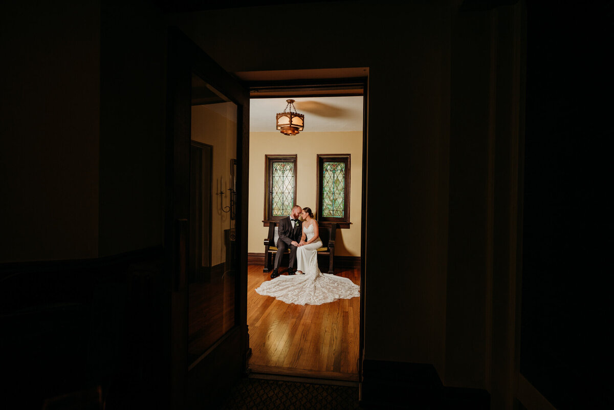 Photo of a bride and groom sitting on a bench and cuddling while being framed by a doorway