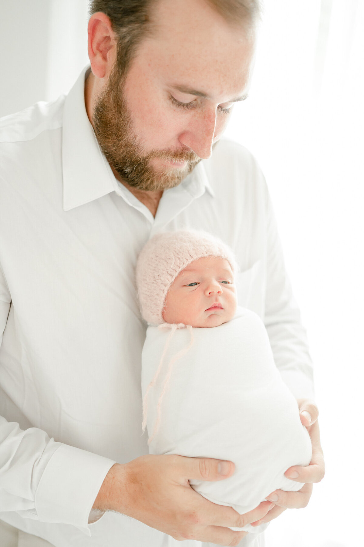 newborn in a pink bonnet is held by her father In Kristie Lloyd’s Nashville newborn photographer studio