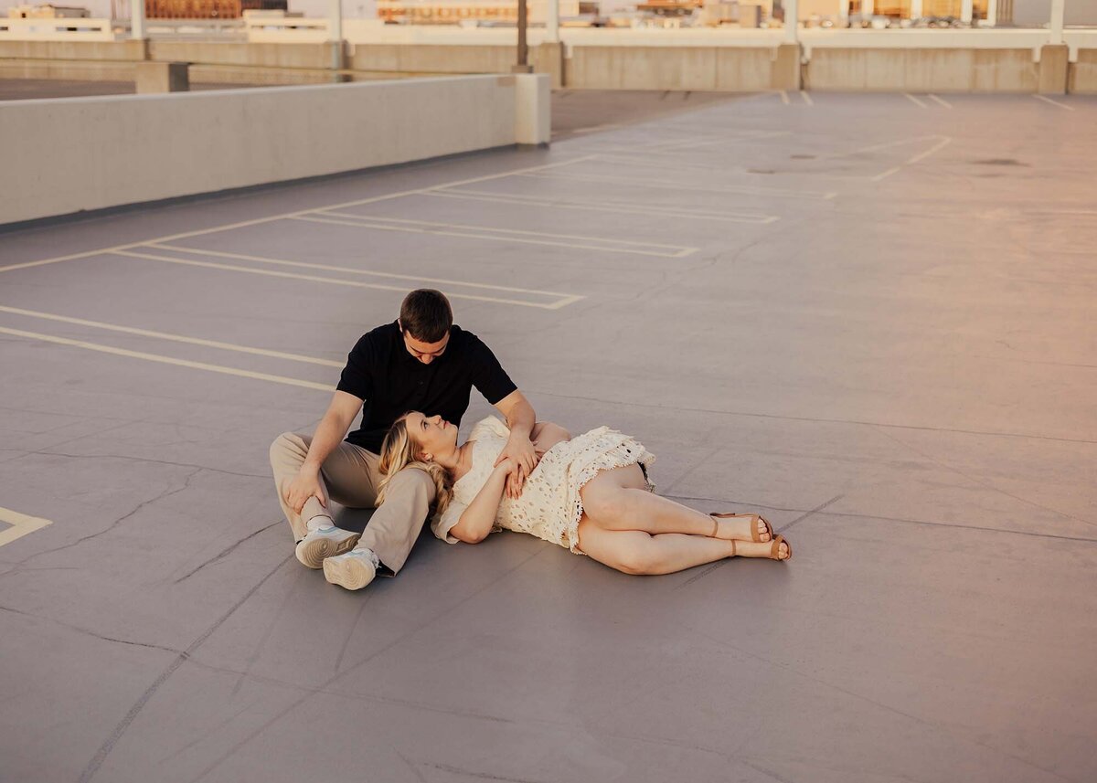 Maddie Rae Photography couple on top of a parking garage. he is sitting and she is laying on one of his legs looking up at him. this is a wide angle shot