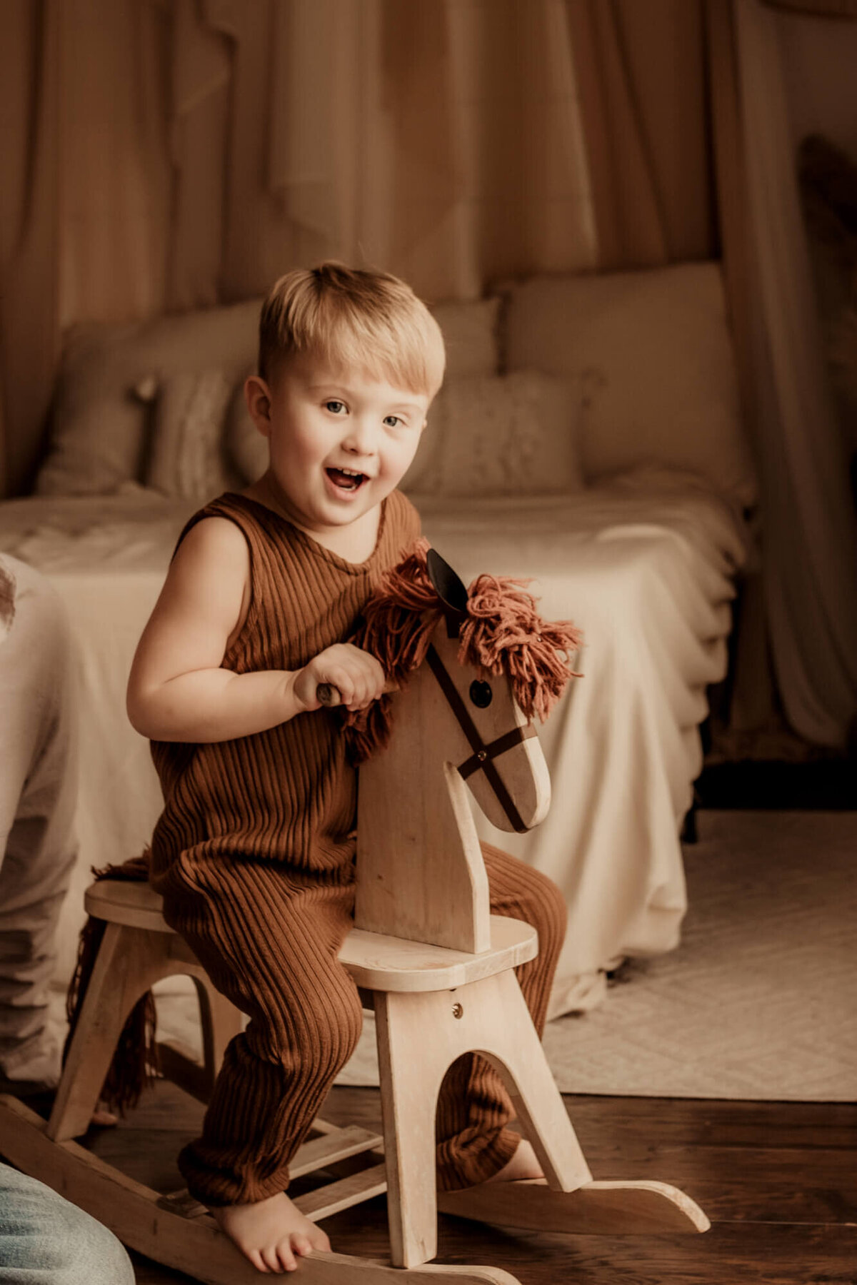 Little boy wearing a brown romper smiles while playing on a rocking horse.