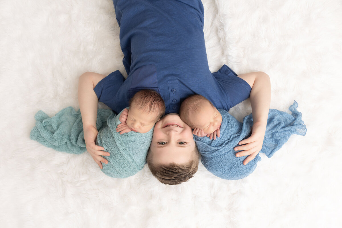 A toddler boy in a blue shirt smiles while laying with his newborn twin siblings sleeping on his shoulders