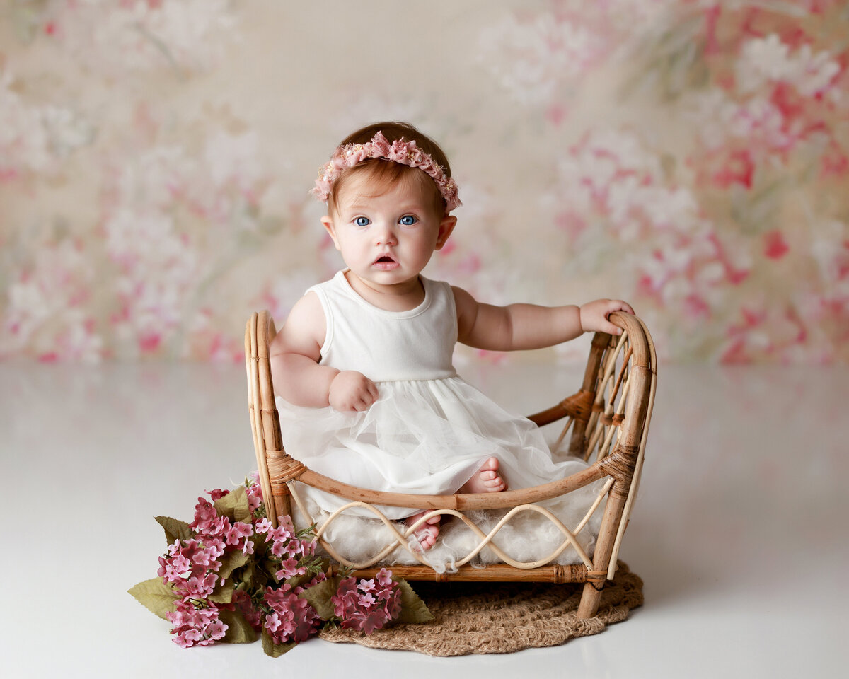 Baby girl 12-month milestone session at top West Palm Beach and Jupiter photography studio. Baby is sitting in miniature rattan photography prop crib in a white dress looking at the camera. Baby has big blue eyes and a floral headband. In the background there is a floral patterned backdrop with shades of gold, pink, and cream.