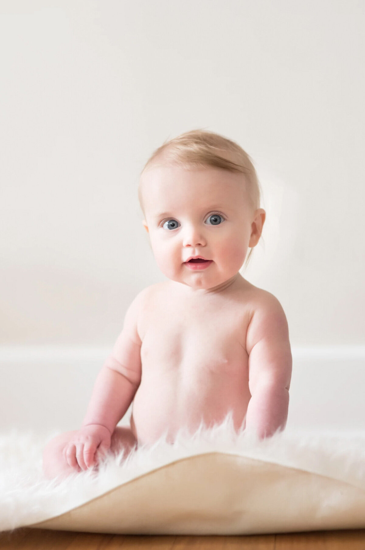 Toddler boy, shirtless, sitting on a fuzzy white blanket in front of a cream colored wall