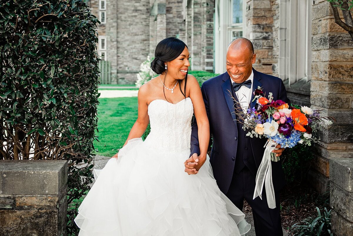 The bride and groom sit on a bench outside Scarritt Bennett stone church. They are holding hands and laughing. The bride has a strapless dress with a sweetheart neckline and a ruffled organza skirt. The groom is wearing a black tuxedo with a black bow tie and a white shirt. He is holding the bride's bouquet. The bouquet is deep orange, peep purple, light blue, yellow, ivory and peach. The bouquet is wrapped in a sheer white ribbon with long tails that come off the stems.