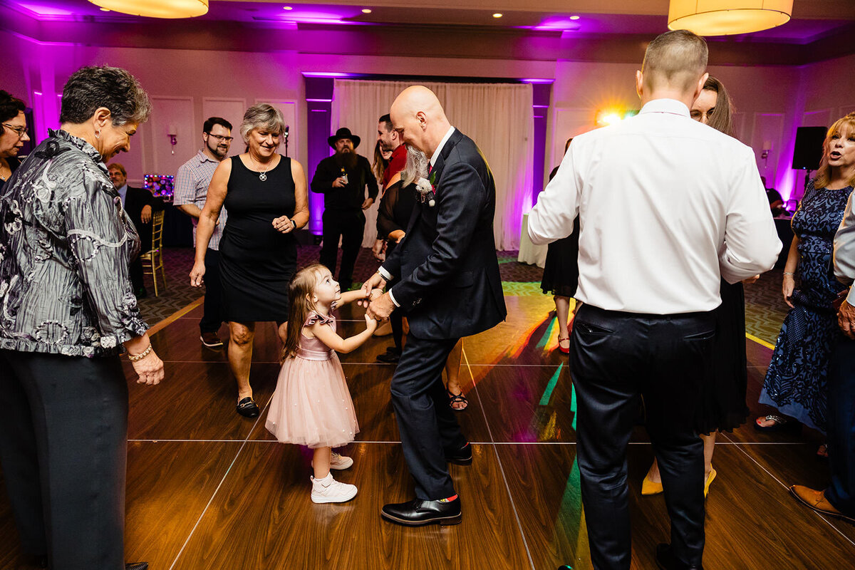 A groom dances with a young girl in a pink dress on the dance floor, surrounded by wedding guests