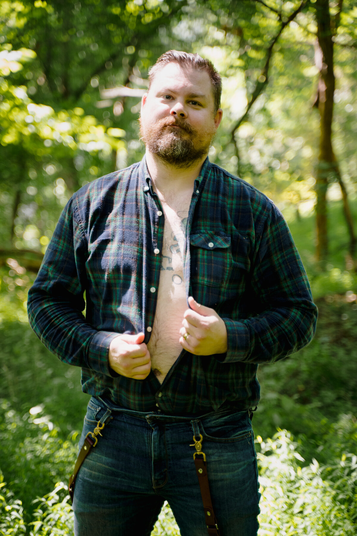 A man standing in the woods in a flannel shirt as he unbuttons it.