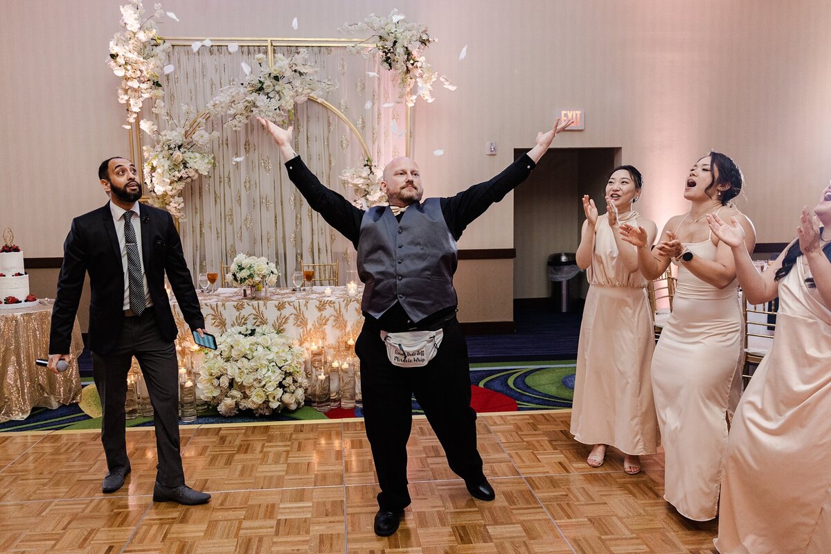 A candid shot of a man pulling out flowers petals from a fanny pack and throwing them into the air at a wedding reception in Dallas, Texas. The man has another guest on his right and three members of the wedding party on his right who all look surprised and excited by his actions. Behind him is the head table adorned with many floral arrangements.