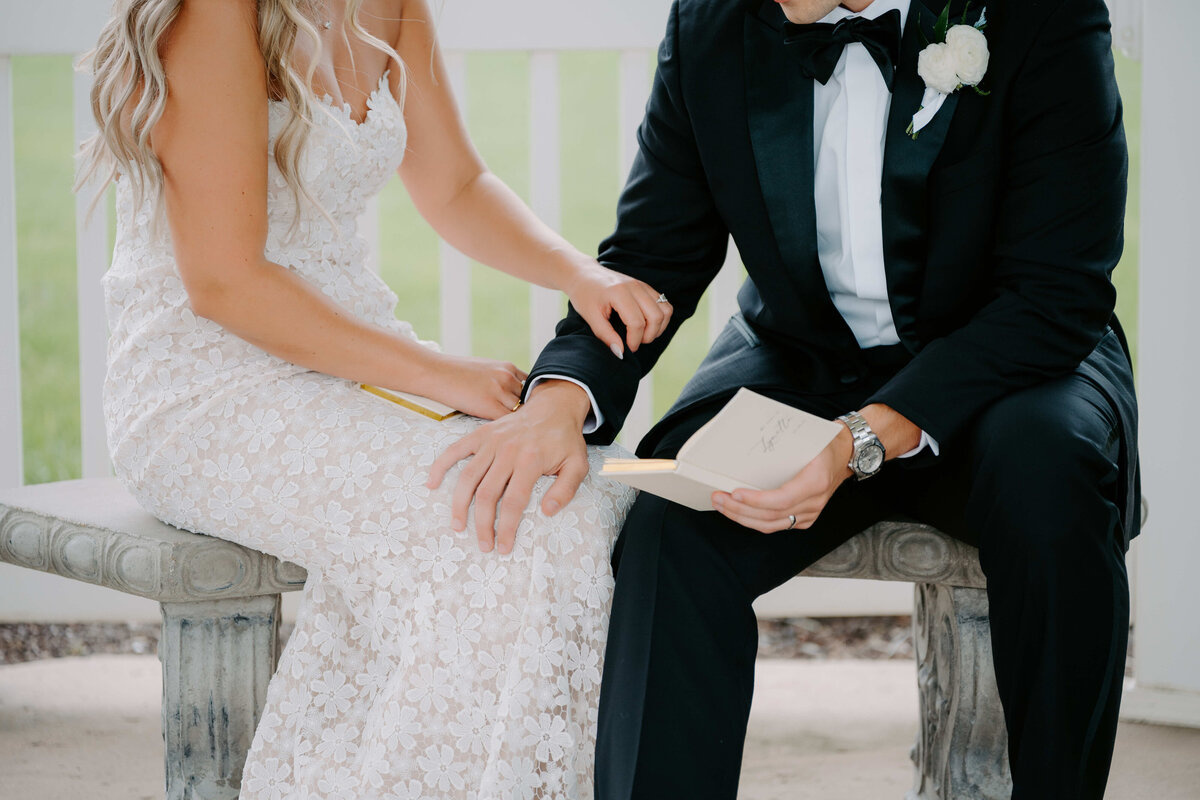 A bride in a lace dress and groom in a tuxedo sit closely on a bench, holding hands and a letter. The scene is intimate and warm, conveying love.