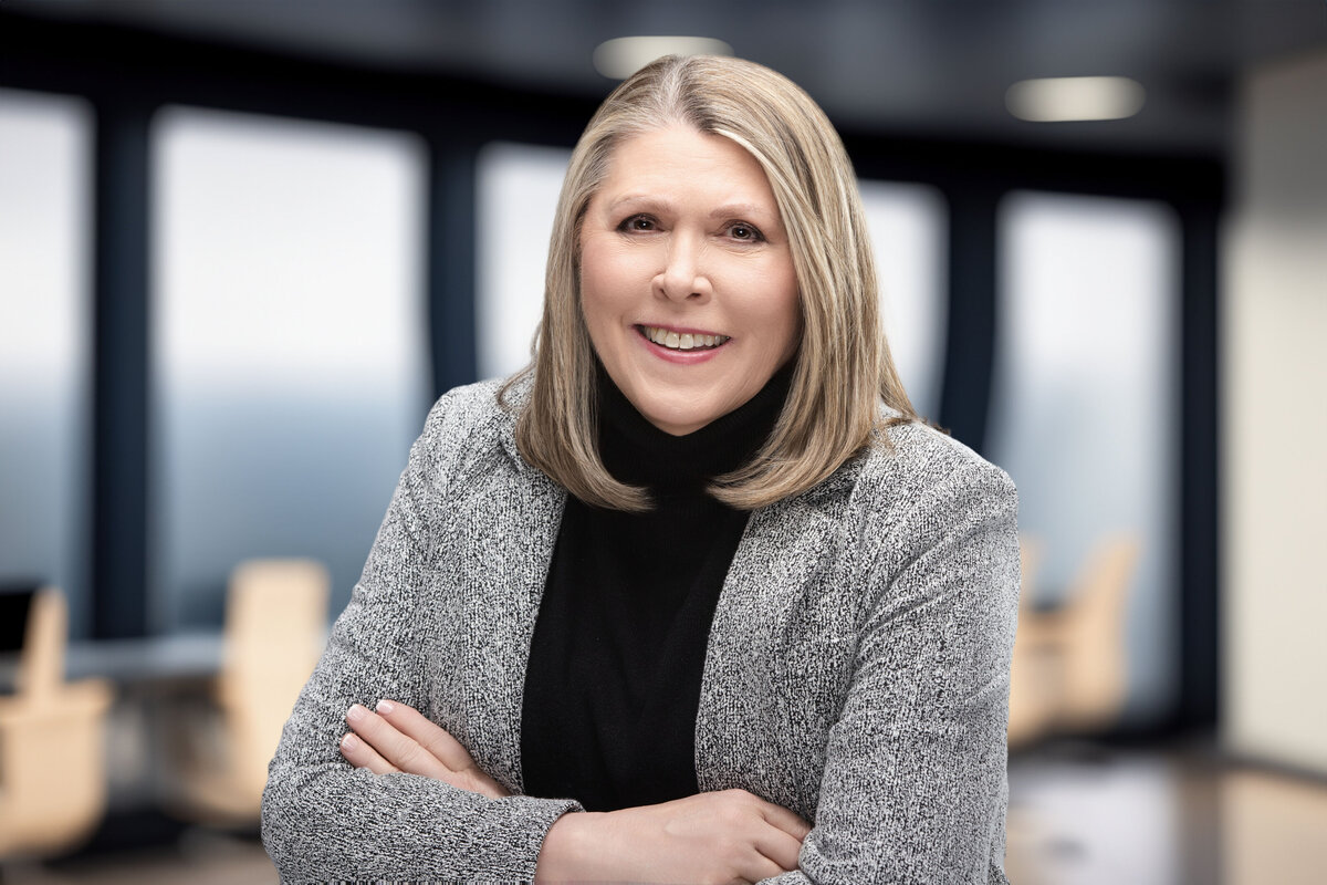 Corporate headshot of a mature professional woman with straight blonde hair and a warm, confident smile, wearing a textured gray blazer and black turtleneck. Captured at Janel Lee Photography, a Cincinnati-based headshot and personal branding studio, this photo features a modern, softly lit office background—ideal for LinkedIn profile photos, executive portraits, and business website bios in the Cincinnati, Ohio and Northern Kentucky area.