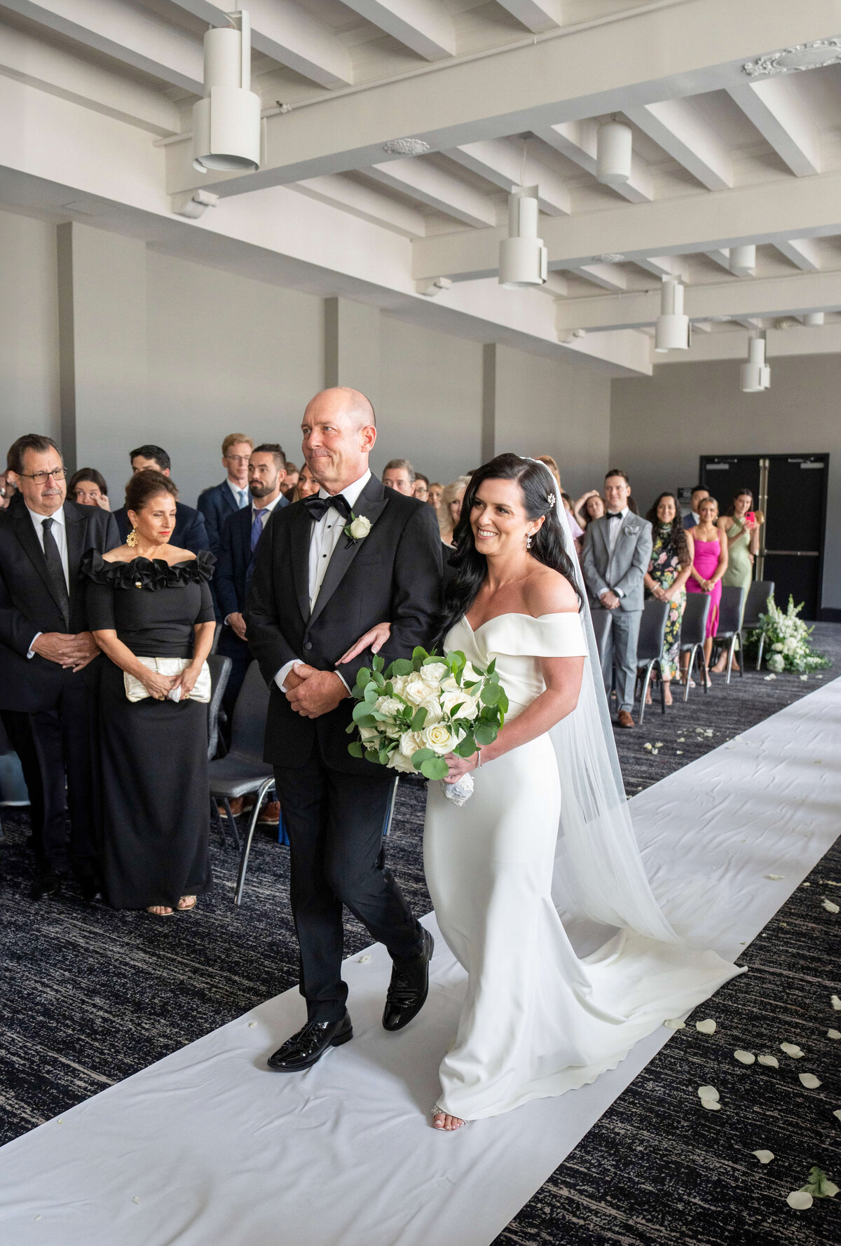 A bride in a white dress and veil walks down the aisle with her father in a tuxedo. Shes holding a bouquet of white flowers. Guests are seated and standing, watching the procession in a decorated, bright room.